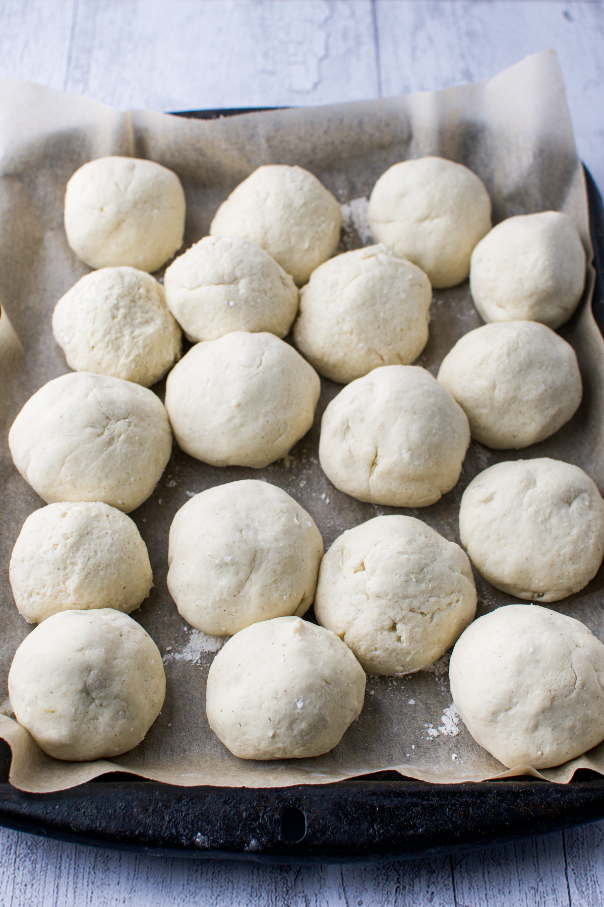 A baking tray lined with parchment paper holds several evenly spaced balls of raw dough, ready for baking. The background shows a lightly textured, white-washed surface.