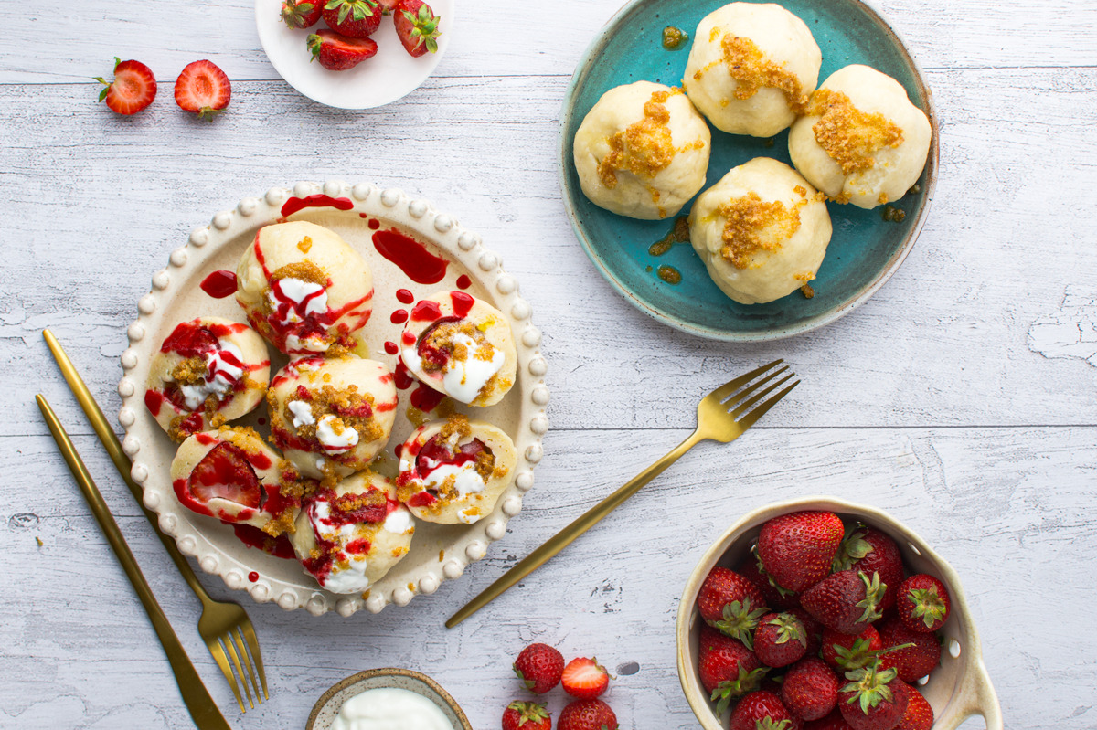 Plates of strawberry dumplings topped with cream and crumbs, whole and sliced strawberries, a small bowl of yogurt, and gold utensils on a white wooden table.
