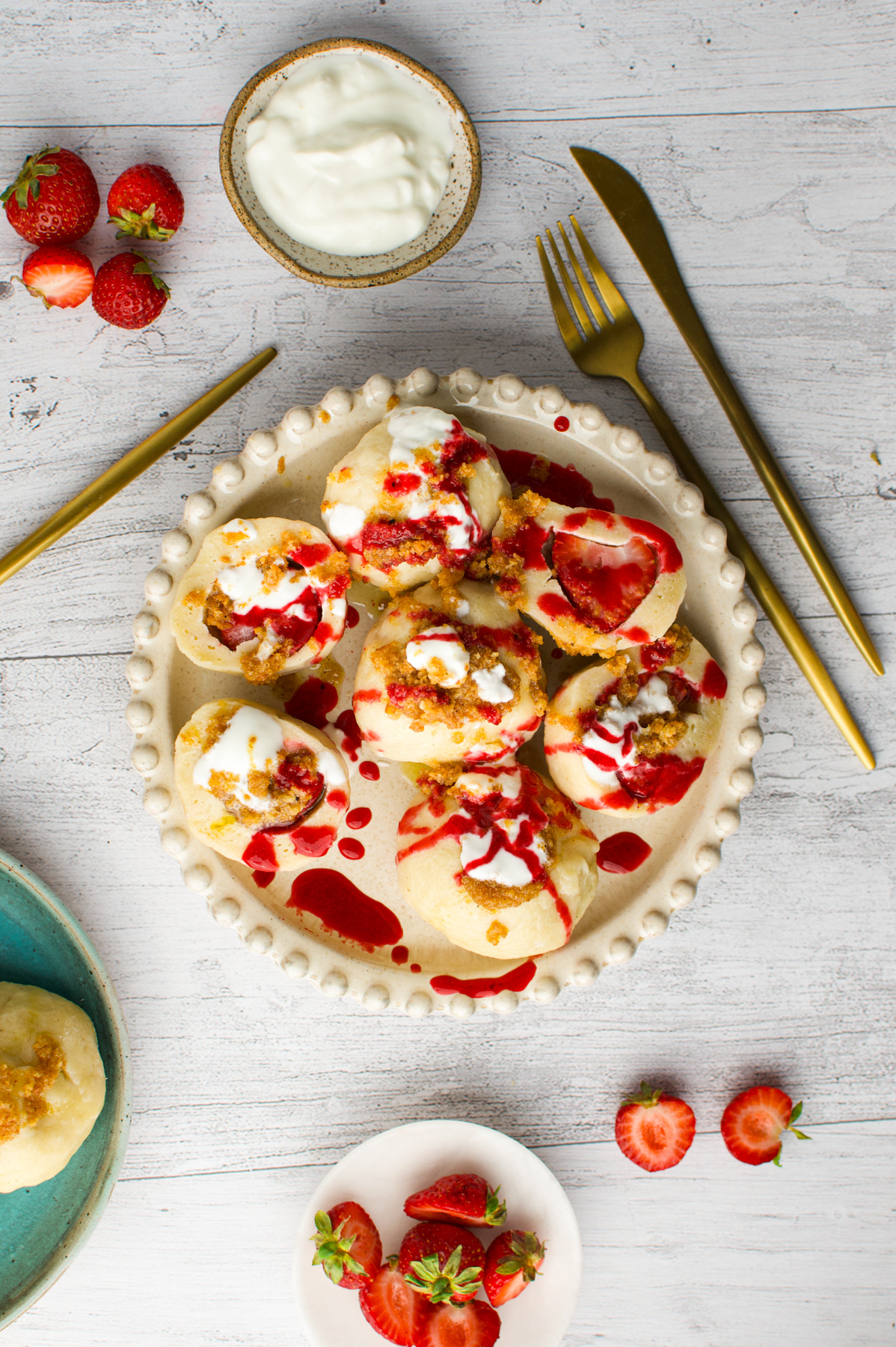 A plate of dumplings topped with white cream and red strawberry sauce, surrounded by fresh strawberries, a bowl of cream, and gold cutlery on a white wooden table.