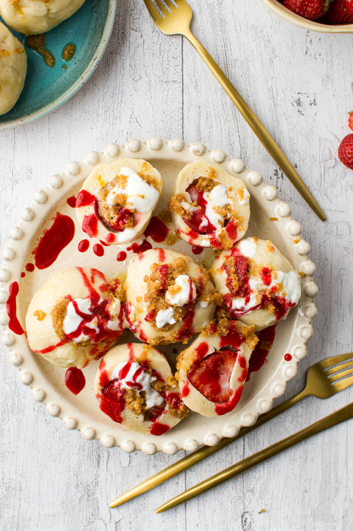 A plate of Polish Strawberry Knedle (Potato Dumplings Gluten-Free) topped with white cream and red fruit sauce, garnished with brown crumbs, sits on a white wooden table with gold cutlery and a small plate of strawberries nearby.