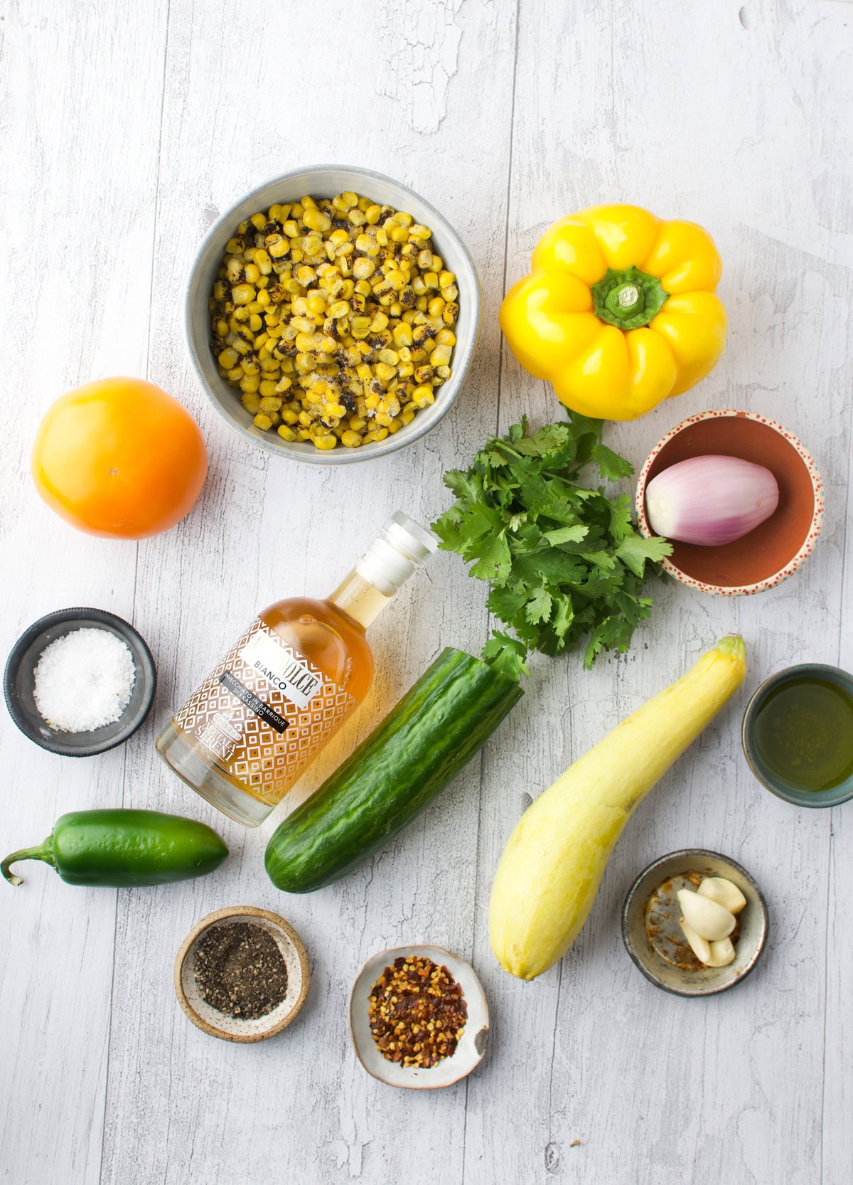 A flat lay of fresh ingredients on a white wooden surface, including yellow squash, cucumber, yellow bell pepper, cilantro, shallot, tomato, jalape&ntilde;o, garlic, corn, oil, vinegar, salt, pepper, and chili flakes.