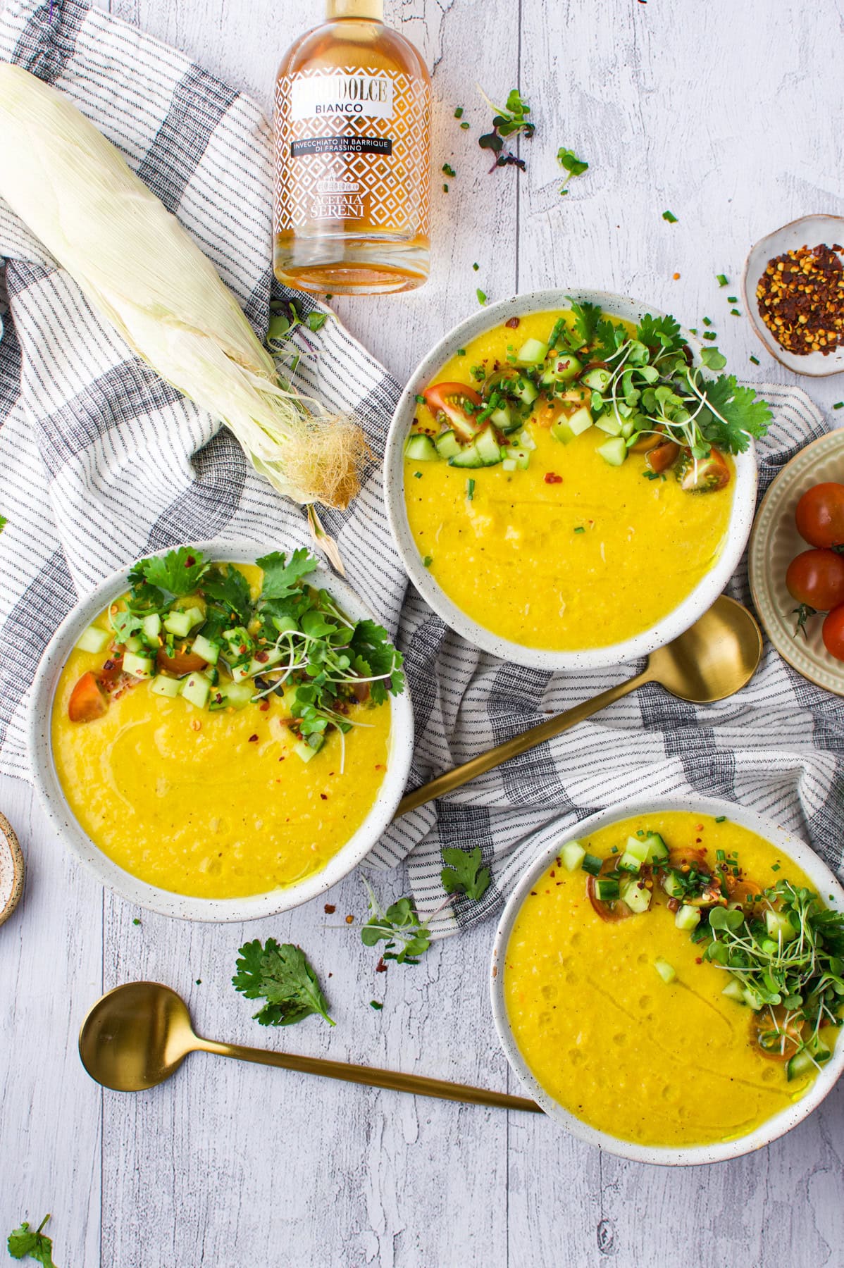 Three bowls of yellow soup topped with diced cucumber, tomato, cilantro, and sprouts on a striped towel, with gold spoons, a corn cob, a bottle, and scattered herbs on a white wooden surface.