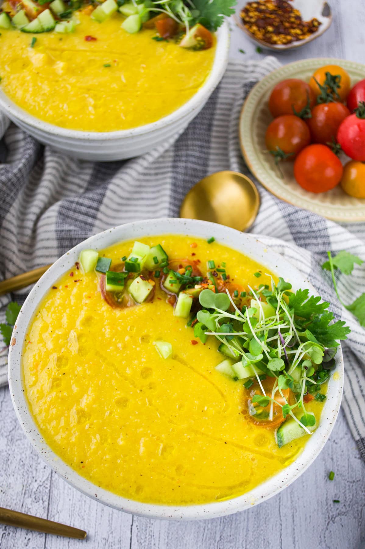 A bowl of bright yellow gazpacho garnished with diced cucumber, microgreens, cilantro, and red pepper flakes sits on a table next to a gold spoon, tomatoes, and a gray-striped cloth.