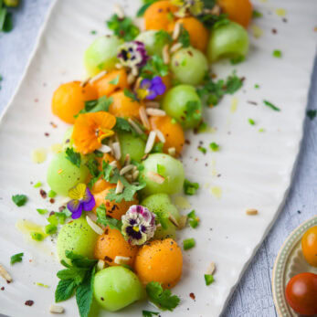 Summer Melon Salad, garnished with edible flowers, herbs, seeds, and cheese shavings, arranged artfully on a light surface.
