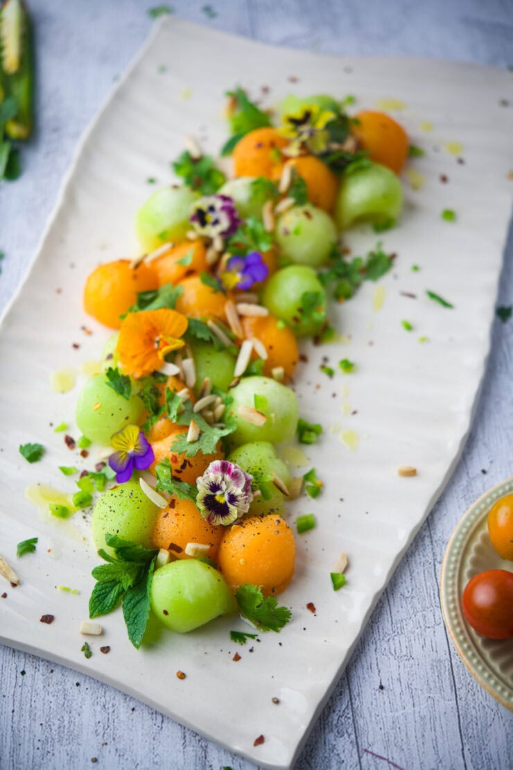 Summer Melon Salad, garnished with edible flowers, herbs, seeds, and cheese shavings, arranged artfully on a light surface.
