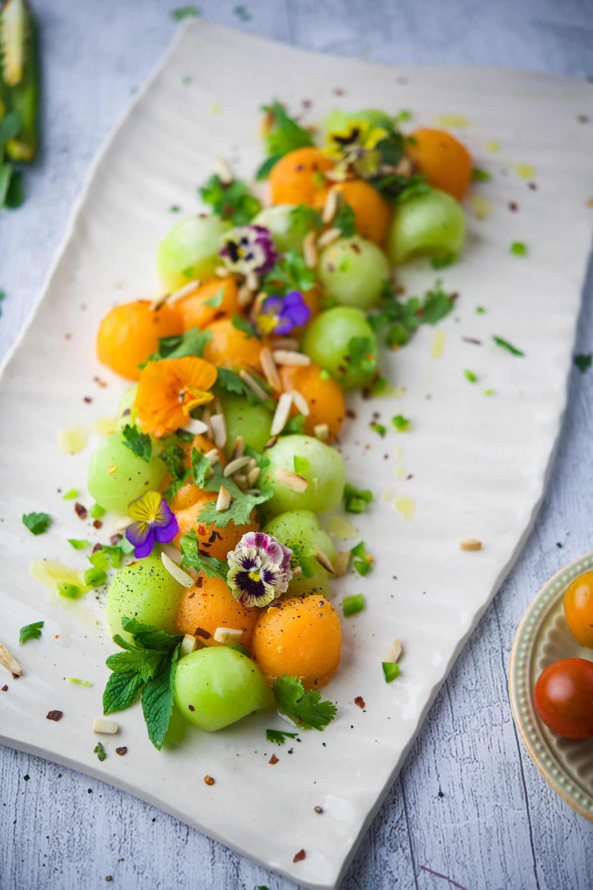 Summer Melon Salad, garnished with edible flowers, herbs, seeds, and cheese shavings, arranged artfully on a light surface.