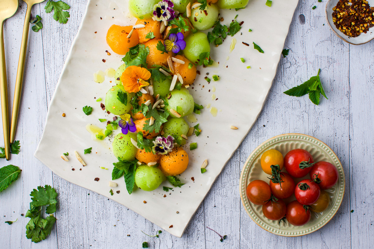 Summer Melon Salad with herbs, edible flowers, and seeds, next to a small bowl of cherry tomatoes on a rustic white table. Gold utensils and scattered herbs surround the dishes.