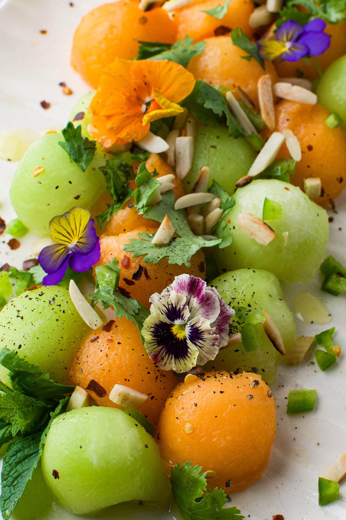 Colorful melon balls of green and orange are garnished with edible flowers, fresh herbs, almond slivers, and cracked black pepper on a white surface.