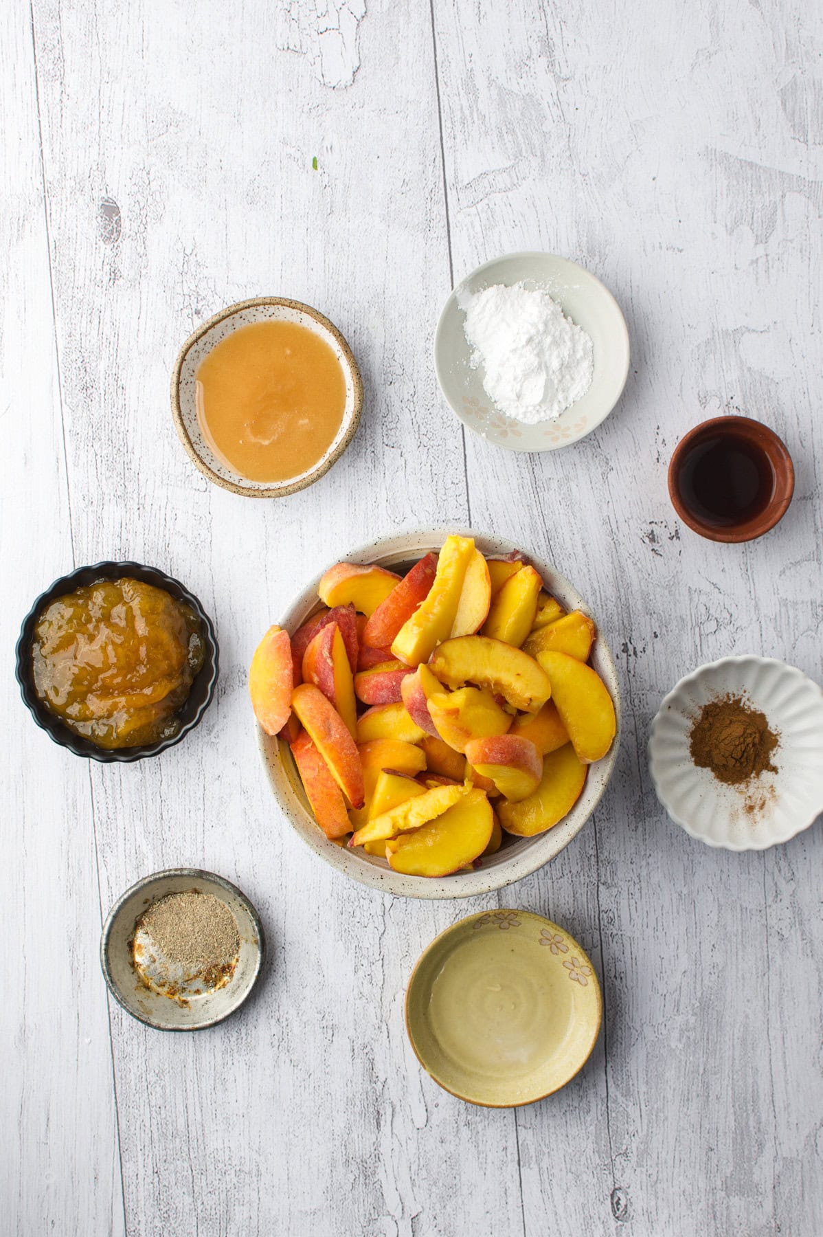 Seven small bowls on a white wooden surface, each containing a different ingredient: sliced peaches, brown sauce, cinnamon, white powder, thick golden sauce, a beige paste, and a light tan powder.