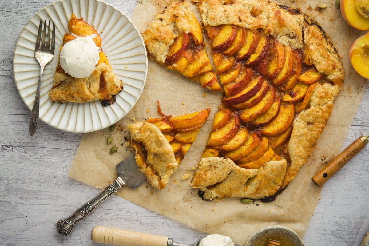 A rustic peach galette with a golden crust sits on parchment paper. A slice has been cut and placed on a plate, topped with a scoop of vanilla ice cream. Fresh peaches and utensils surround the dessert.