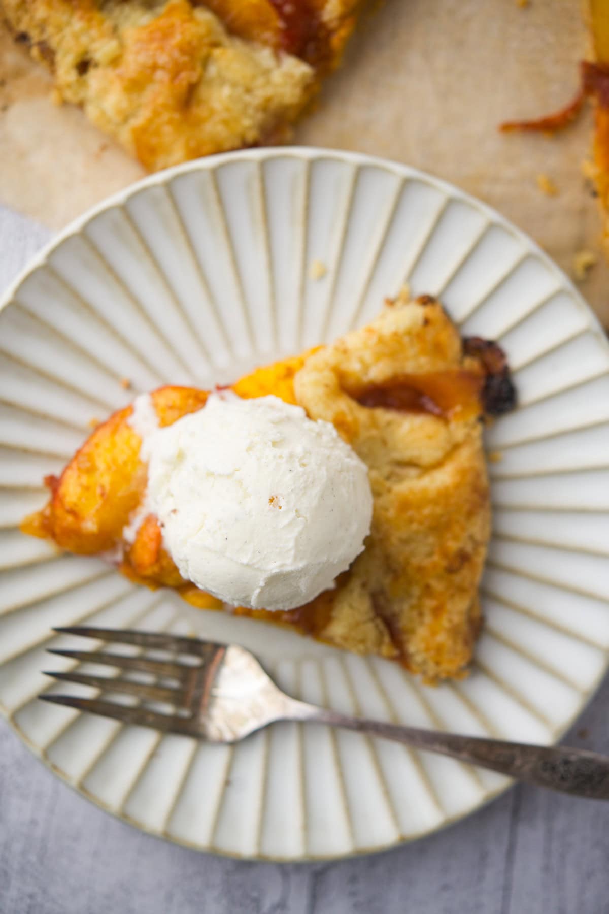 A slice of fruit galette topped with a scoop of vanilla ice cream sits on a white, ridged plate next to a fork.
