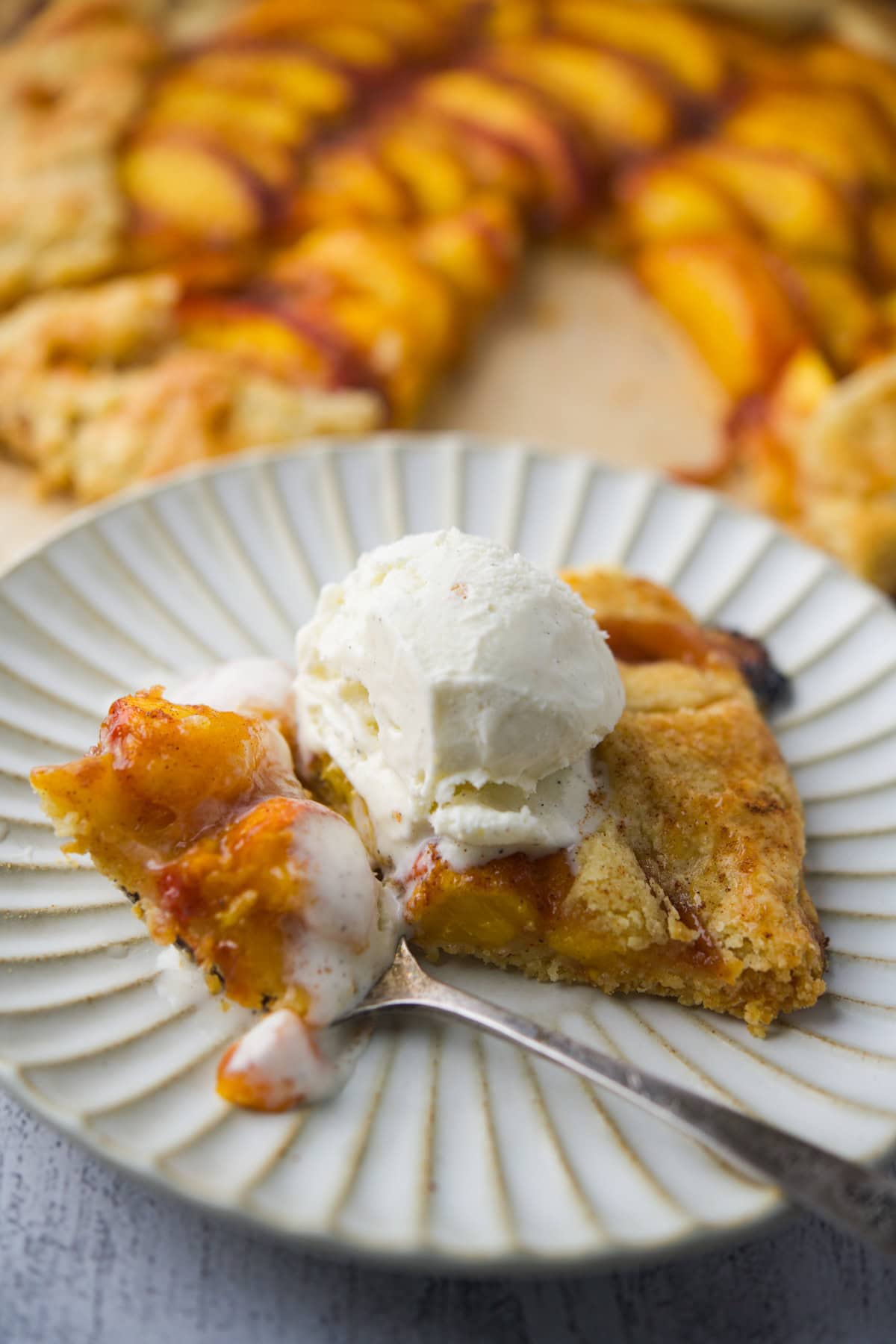 A slice of gluten-free peach galette topped with a scoop of melting vanilla ice cream sits on a plate with a fork, with the rest of the galette blurred in the background.