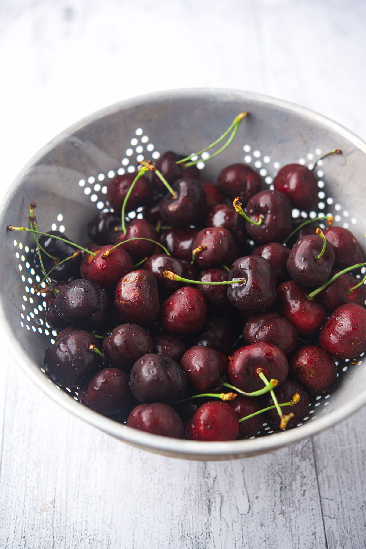 A metal colander filled with fresh, washed cherries, some with stems attached, sits on a light wooden surface. The cherries are deep red and glistening with droplets of water.