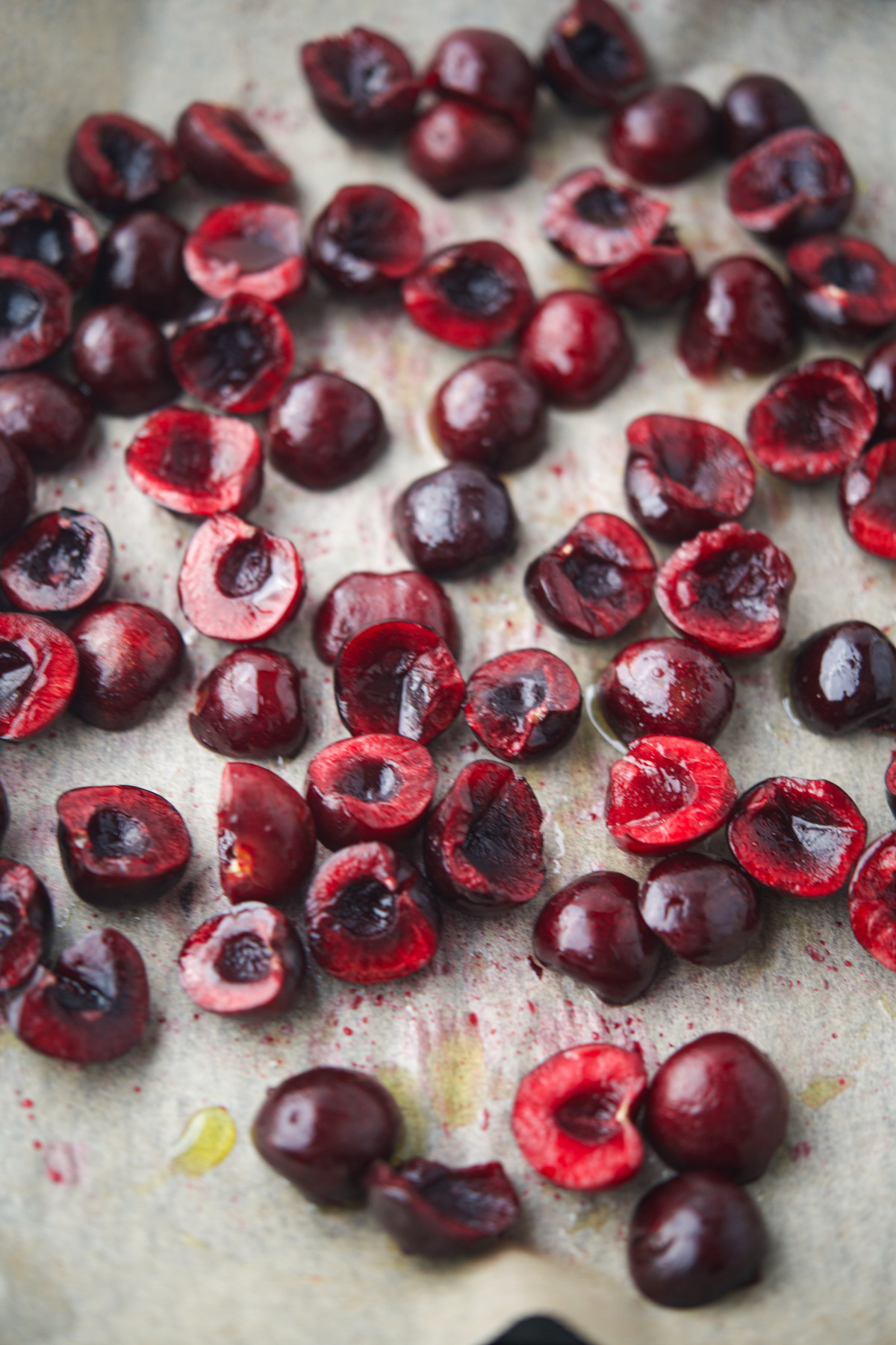 Halved dark red cherries scattered on a sheet of parchment paper, some with pits removed and some with pits intact.