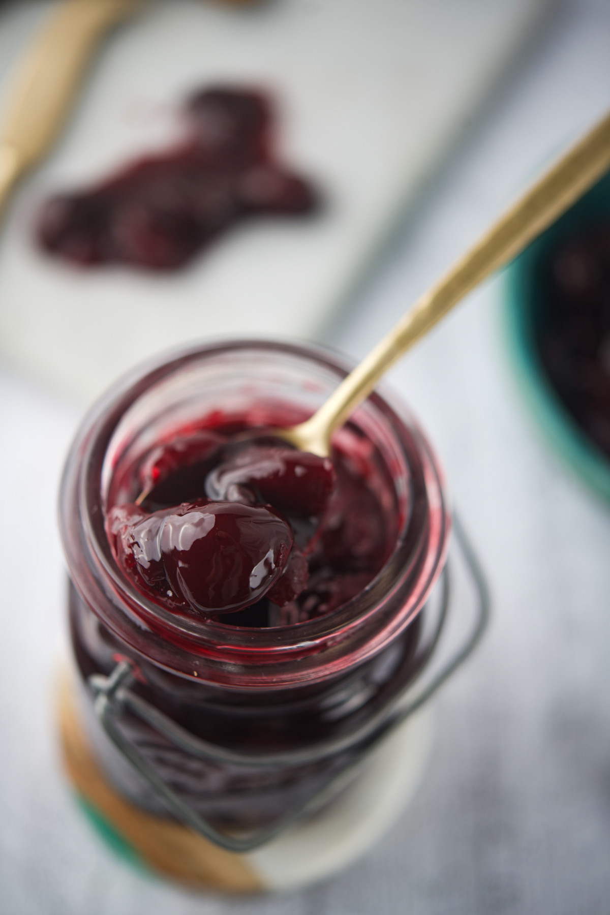 A close-up of a glass jar filled with glossy, dark red cherry preserves. A gold spoon is scooping out some of the preserves, with cherries visible in the thick syrup. The background is softly blurred.