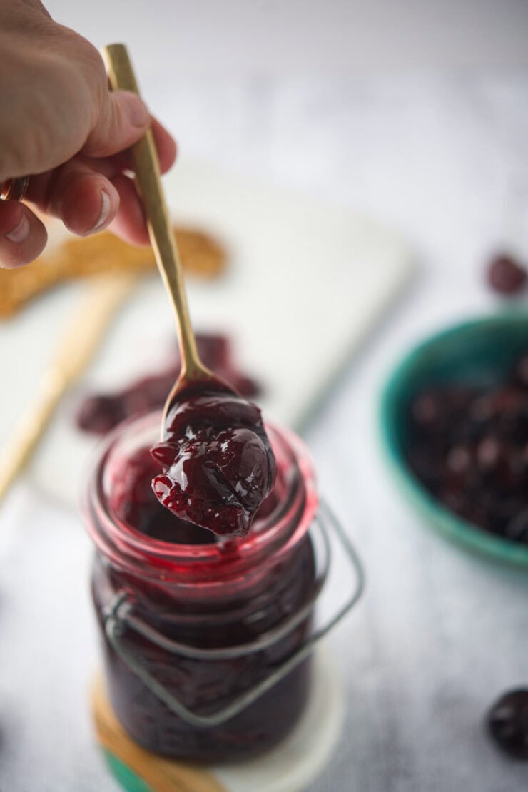A hand holds a spoonful of chunky dark cherry compote above an open glass jar filled with the jam, with a blurred green bowl and cherries in the background.