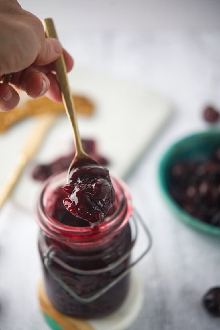 A hand holds a spoonful of chunky dark cherry compote above an open glass jar filled with the jam, with a blurred green bowl and cherries in the background.