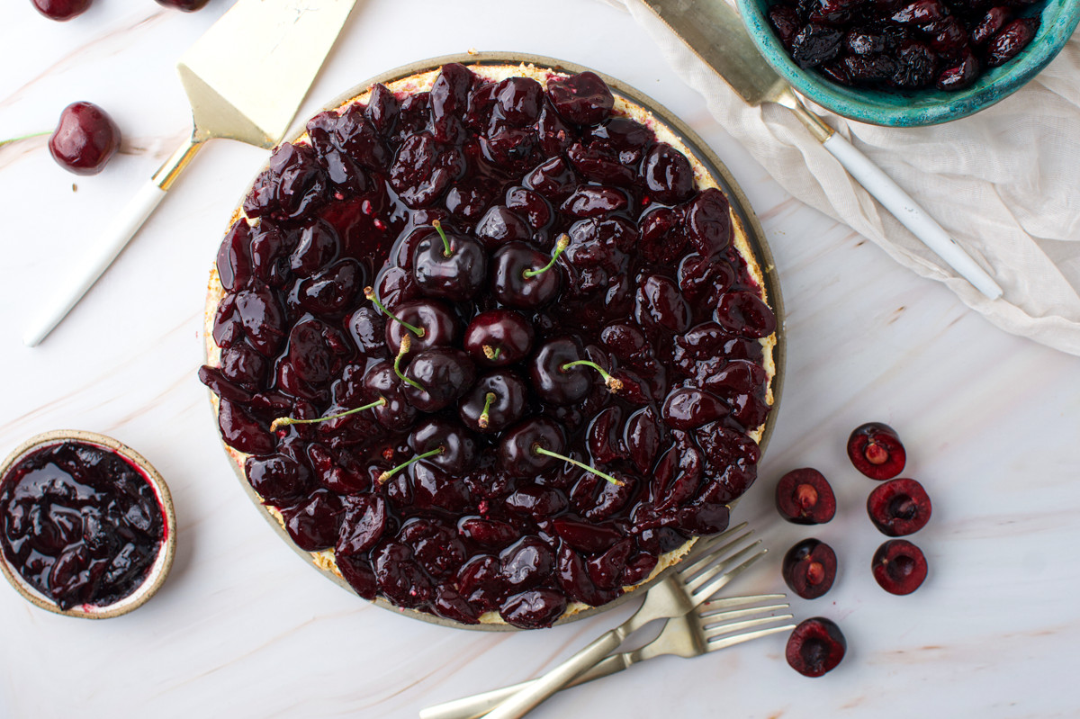 A cherry cheesecake topped with glossy, fresh cherries sits on a white surface. Sliced cherries, forks, and a bowl of cherry topping surround the cake, creating a vibrant, inviting dessert scene.