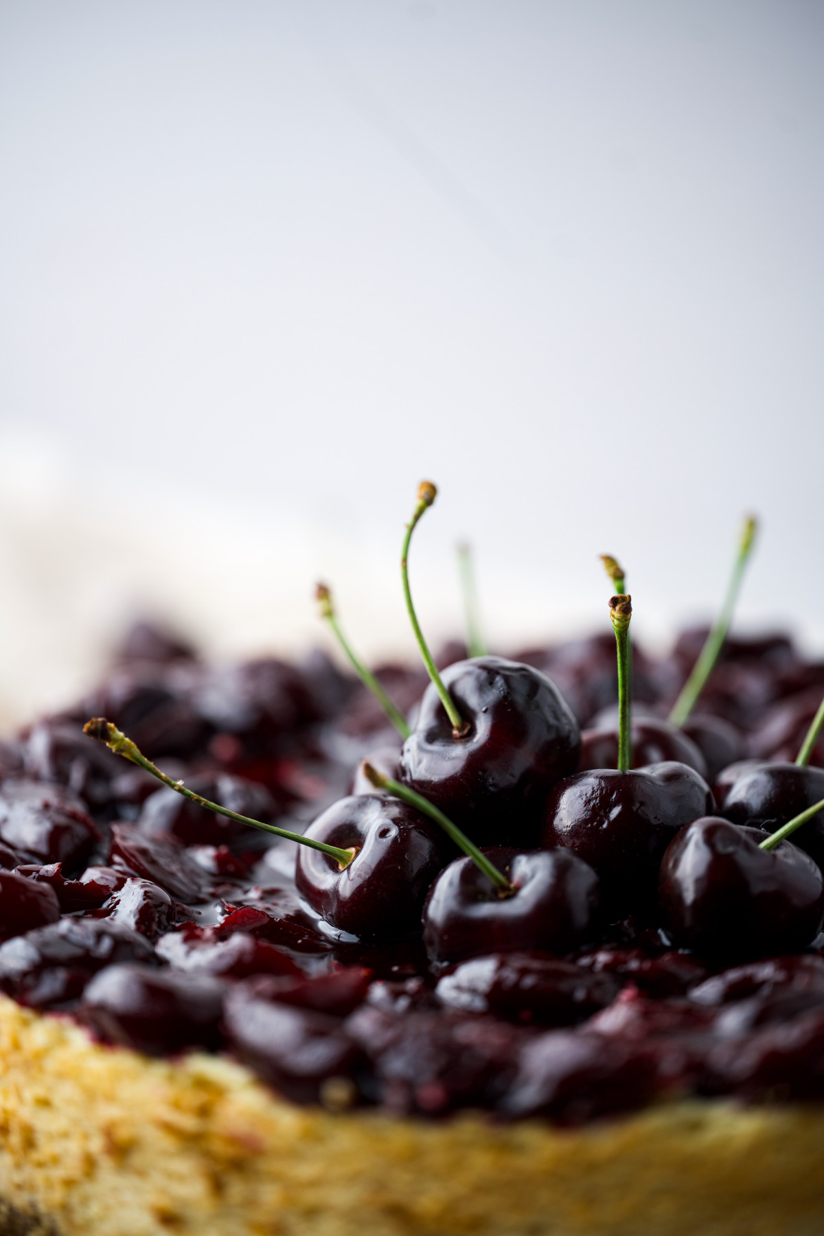 A close-up of a cherry-topped dessert, with glossy, fresh cherries and cherry filling on a cake or tart, set against a light, blurred background.