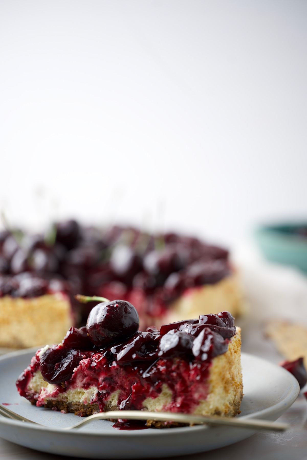 A close-up of a slice of cherry-topped cheesecake on a plate, with the rest of the cheesecake visible in the background. The cheesecake is creamy with a golden crust and glossy cherry topping.