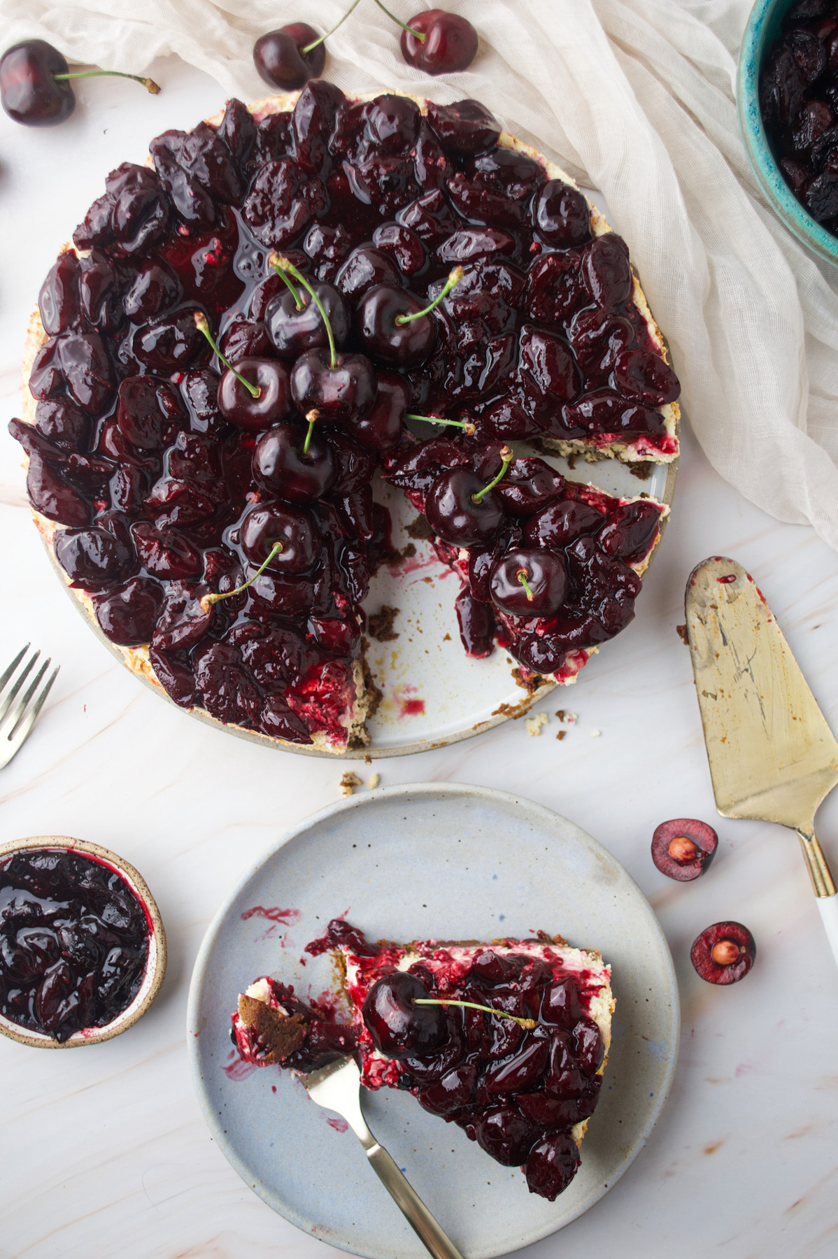 A cherry cheesecake with a glossy, dark cherry topping sits on a white surface. One slice has been served on a gray plate nearby, with scattered cherries and utensils around.