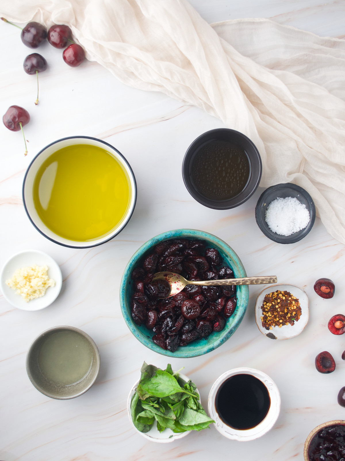 A flat lay of ingredients for Roasted Cherry Vinaigrette on a white surface: a bowl of olive oil, a bowl of black olives with a spoon, salt, sliced cherries, crushed red pepper, minced garlic, basil leaves, balsamic vinegar, and another dark liquid.