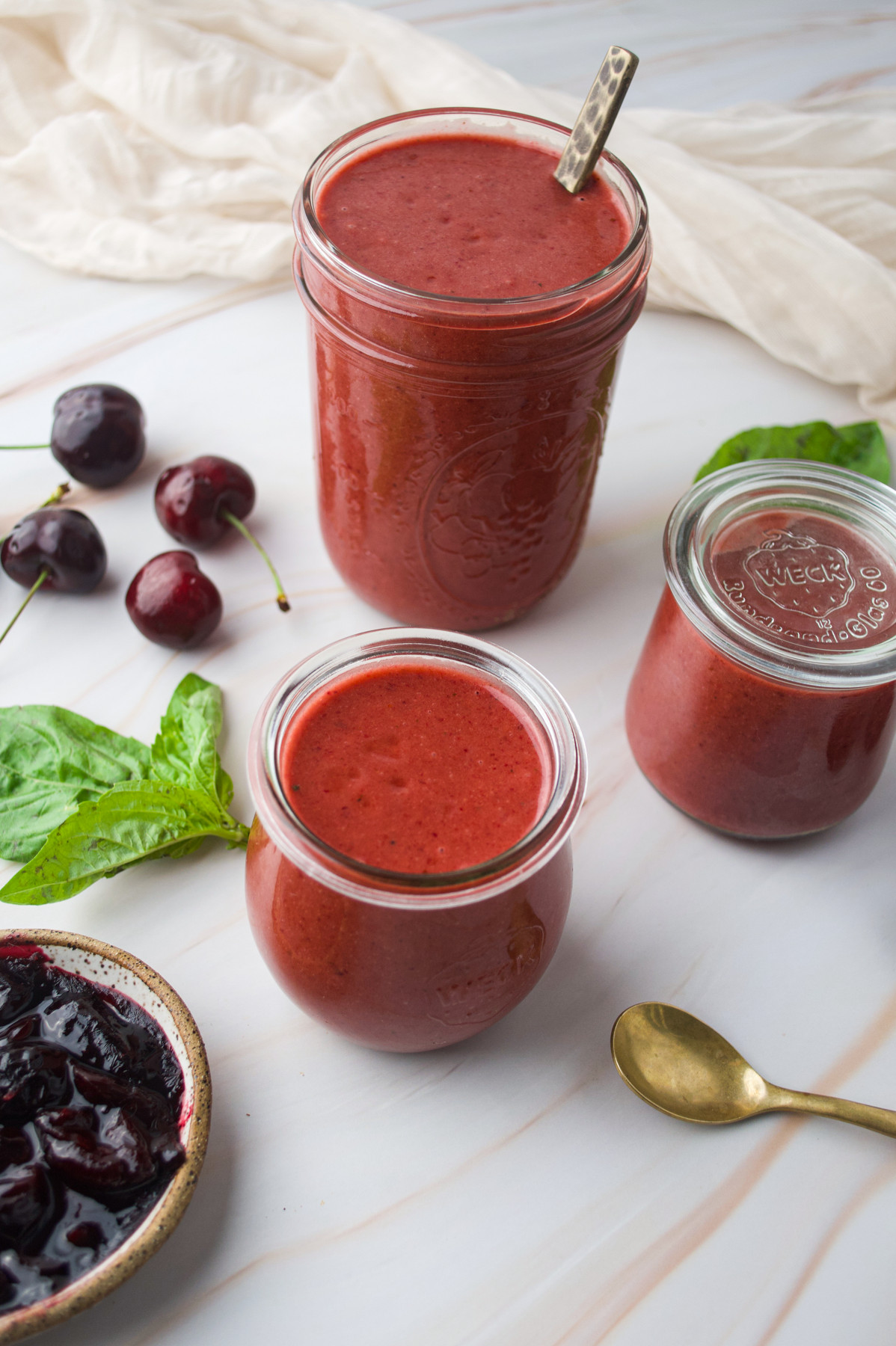 Three jars filled with a thick red smoothie sit on a marble surface, surrounded by fresh cherries, basil leaves, a bowl of cherry jam, a gold spoon, and a light linen cloth in the background.