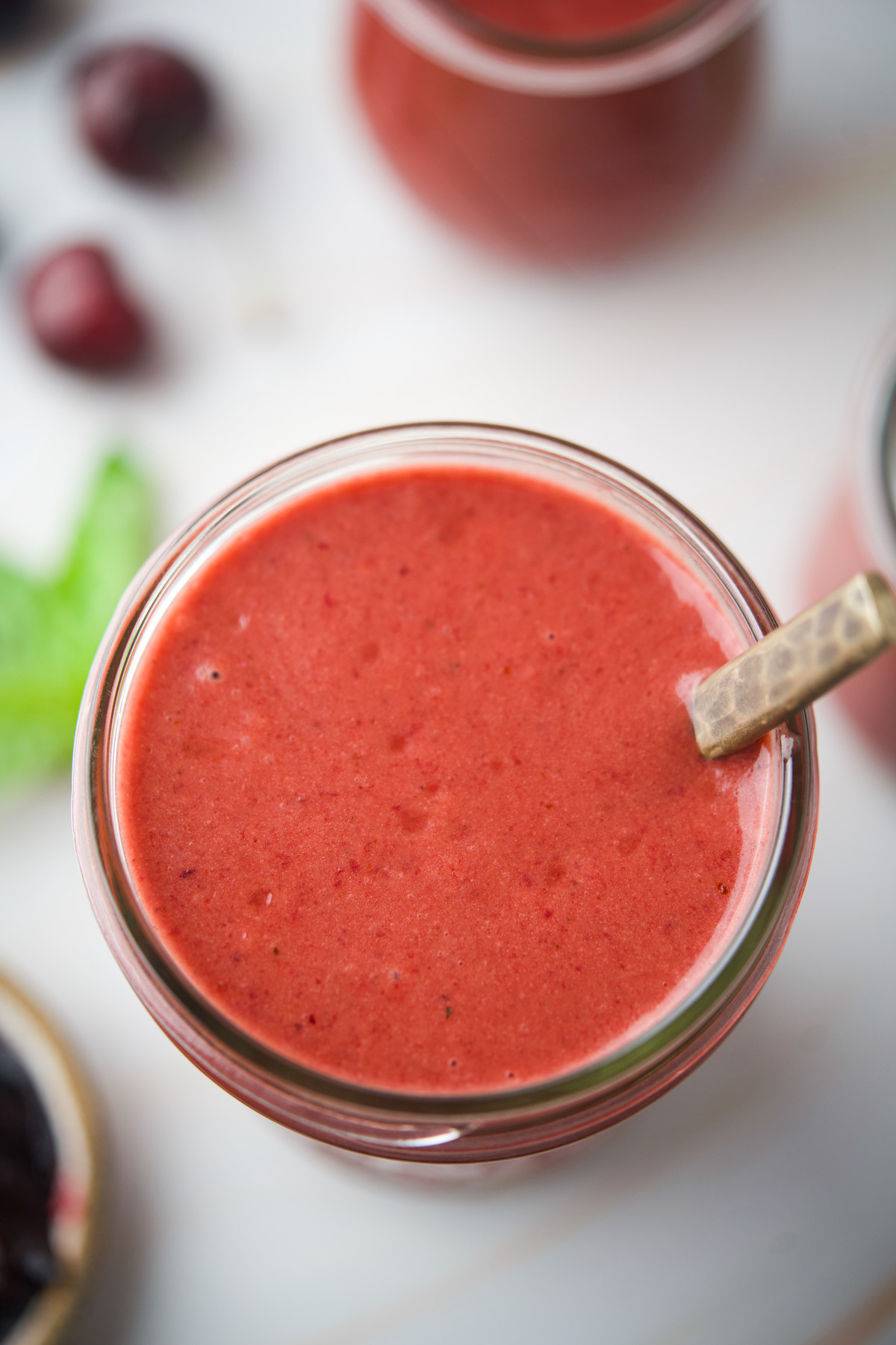 A close-up of a glass filled with Roasted Cherry Vinaigrette with a metal spoon in it. Blurred cherries and green leaves are visible in the background.