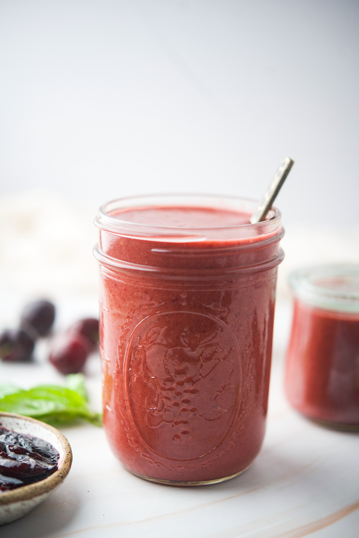 A glass jar filled with a Roasted Cherry Vinaigrette with a spoon inside. In the background, there are blurred plums, a green leaf, and another jar filled with the same smoothie.