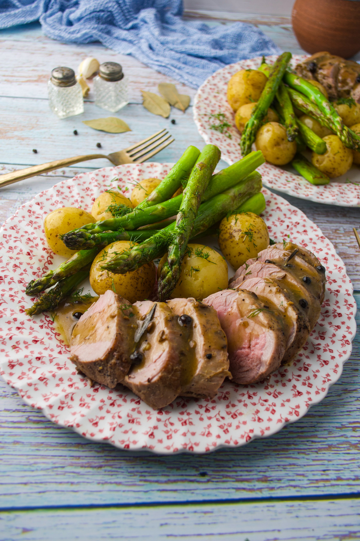 A floral plate with sliced pork tenderloin in sauce, roasted asparagus, and baby potatoes. Another plate with the same food is in the background, on a rustic wooden table with a blue cloth.