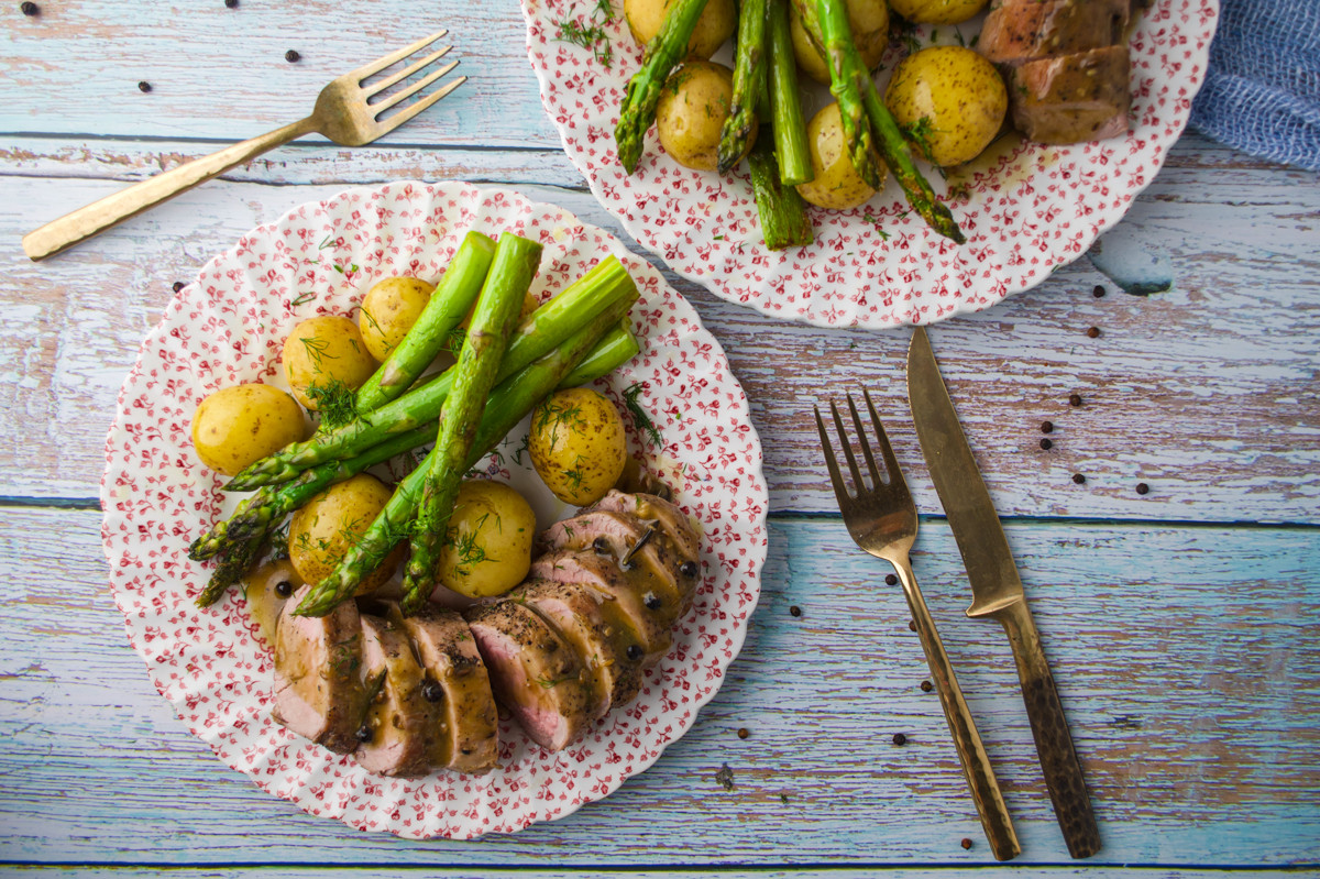 Two floral plates with sliced meat, asparagus, and baby potatoes on a rustic wooden table. Forks and knives are placed beside the plates, peppercorns scattered around.