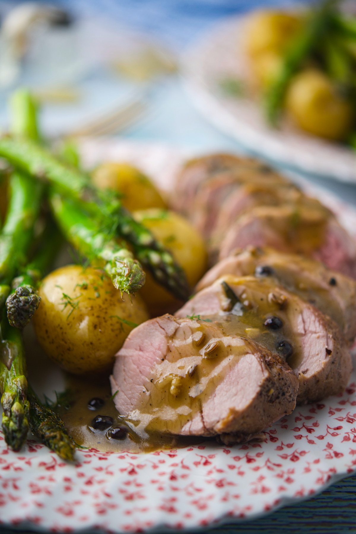 Slices of pork tenderloin topped with sauce, served with roasted baby potatoes and asparagus on a decorative plate.