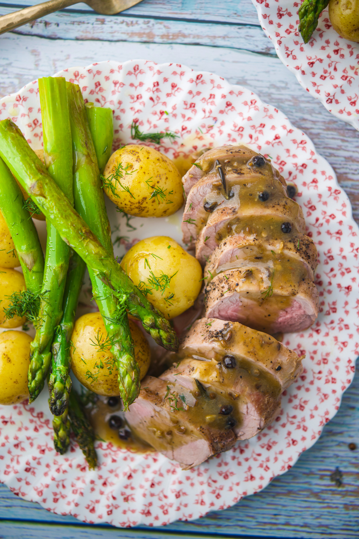 A floral plate with sliced pork tenderloin topped with gravy, whole baby potatoes with dill, and spears of cooked asparagus.