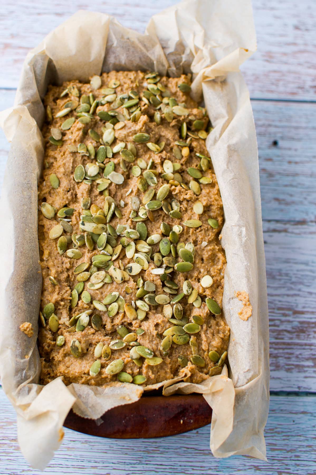 A loaf of bread topped with pumpkin seeds sits in a loaf pan lined with parchment paper on a light wooden surface.