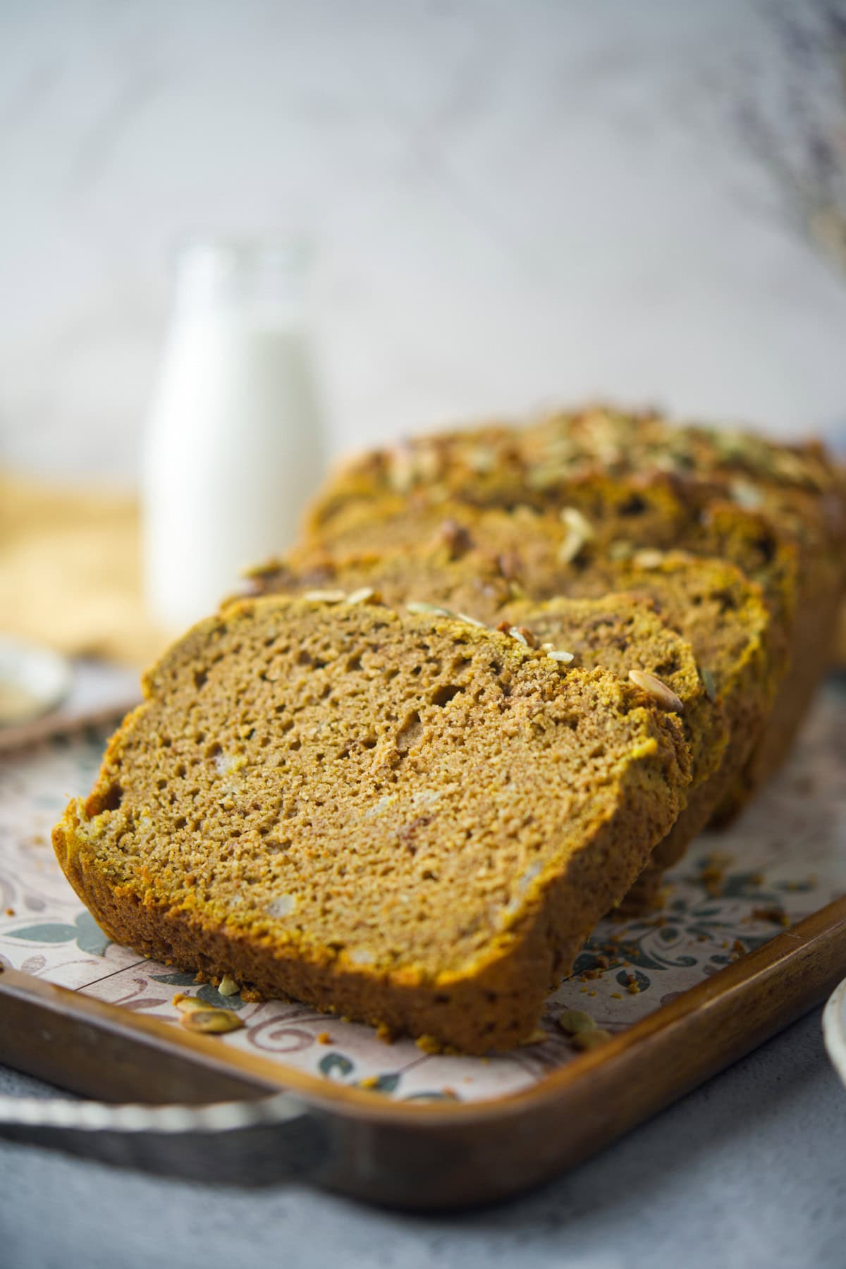 A close-up of sliced pumpkin bread on a decorative tray, with a bottle of milk in the blurred background. The bread appears moist and topped with nuts and seeds.