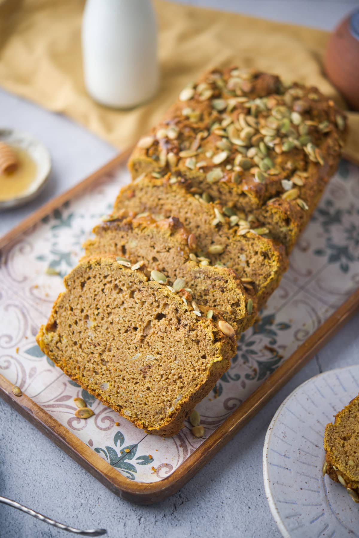 A sliced loaf of gluten-free sourdough pumpkin bread topped with pumpkin seeds sits on a patterned tray. A bottle of milk and a plate with honey and a honey dipper are nearby. The scene is set on a light surface with a yellow cloth in the background.