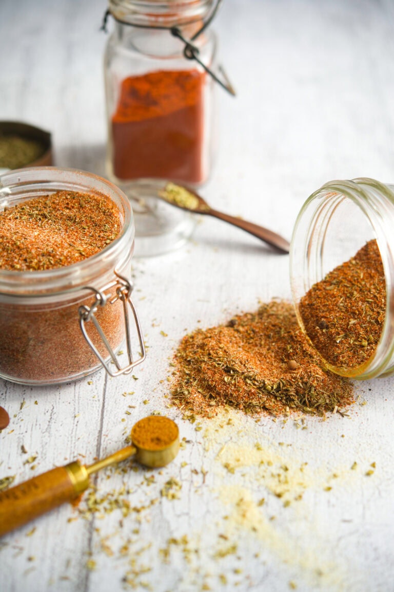Overhead view of homemade Creole spice mix in a glass jar, with some spilled onto a white wooden surface. Surrounding the jar are measuring spoons with paprika, oregano, and garlic powder, showcasing the vibrant, textured blend of herbs and spices.
