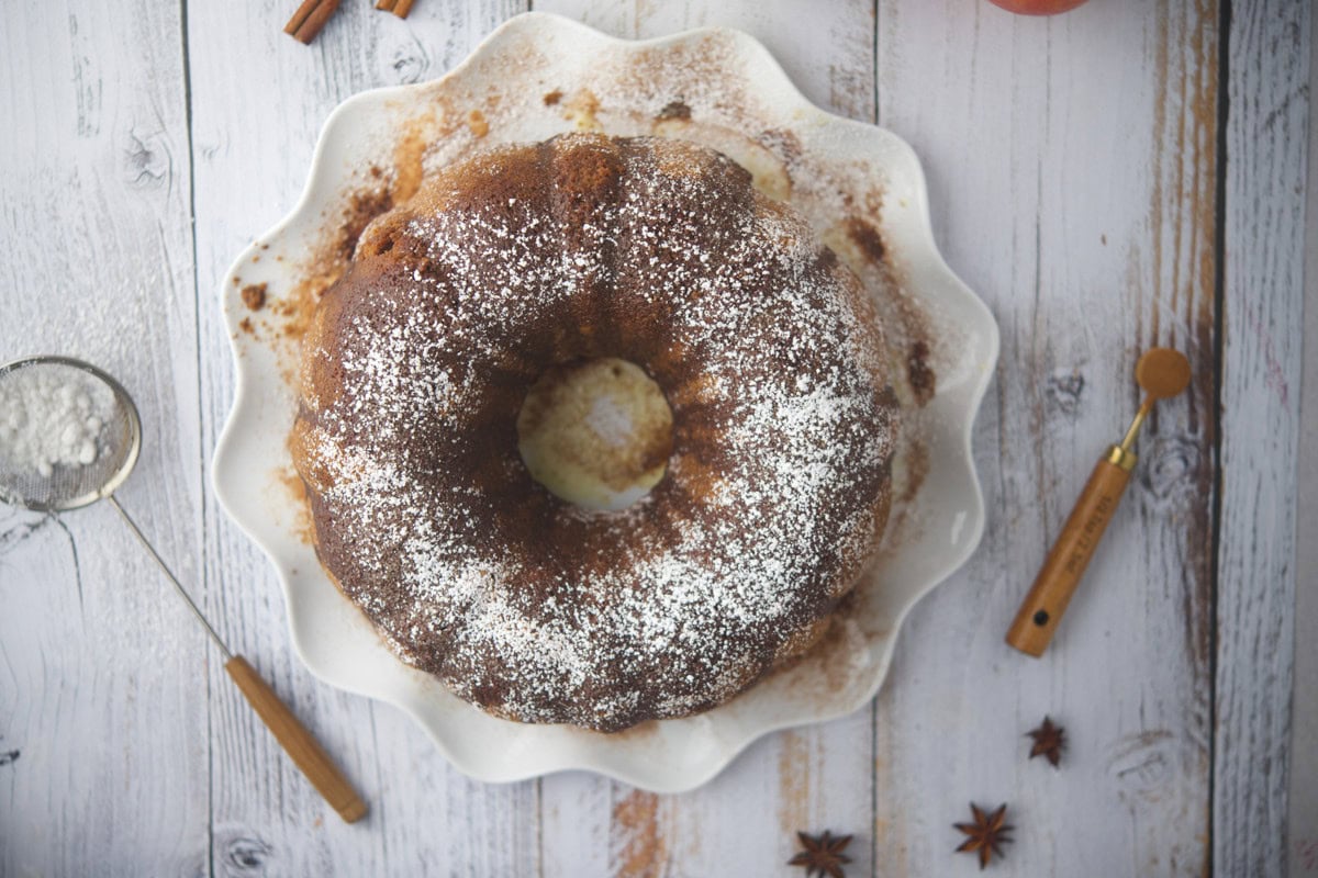 A gluten-free apple cider donut cake dusted with powdered sugar sits on a white scalloped plate atop a rustic wooden table, surrounded by baking tools and spices.