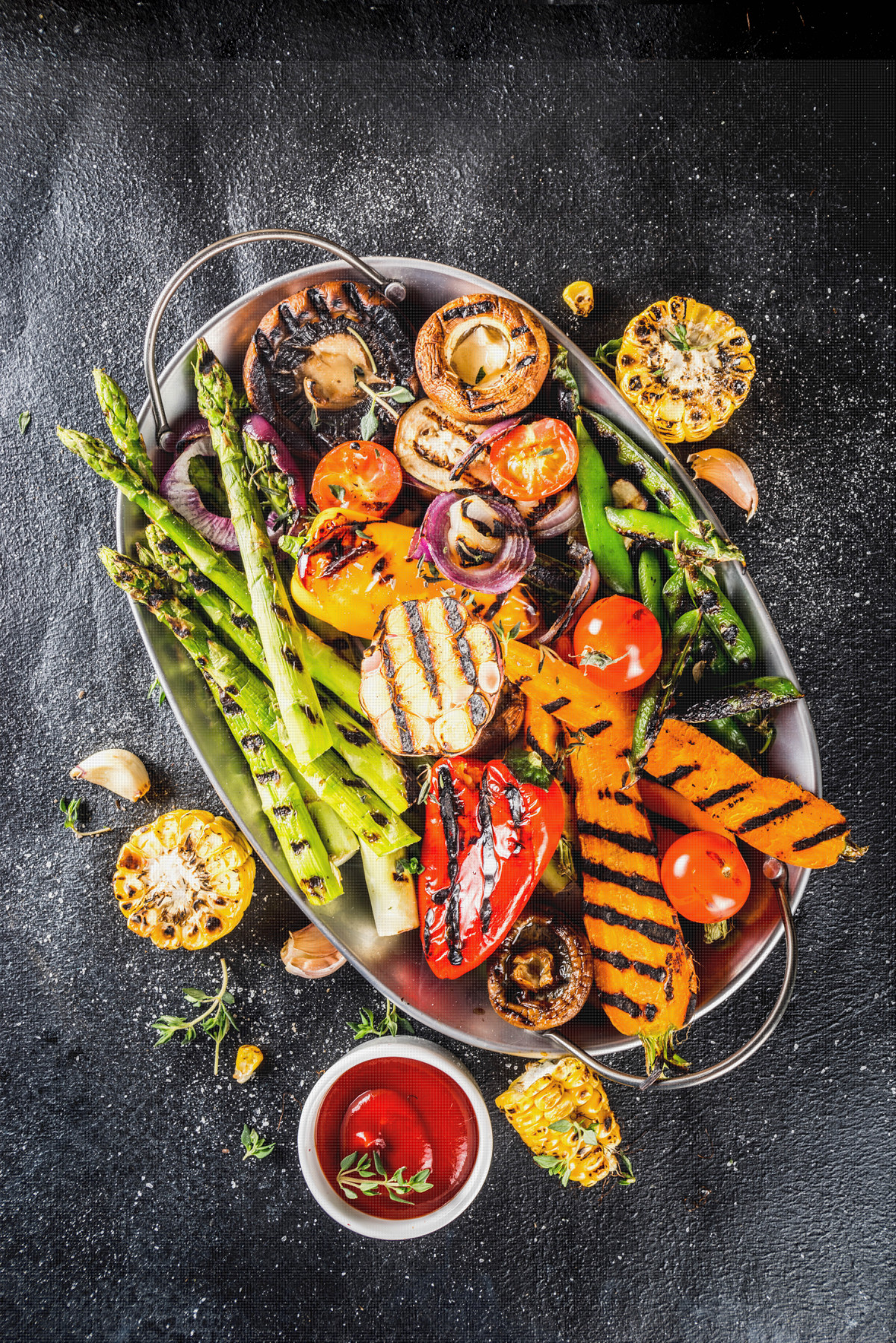 Oval platter with assorted grilled vegetables, including asparagus, mushrooms, corn, peppers, tomatoes, and squash, garnished with herbs. Small bowl of red dipping sauce on the side. Black textured background.