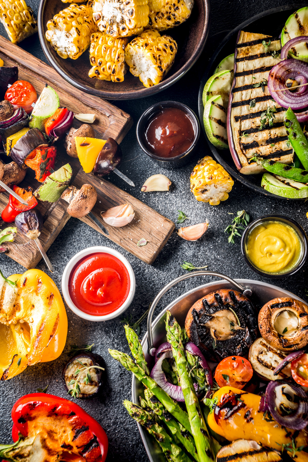 Colorful grilled vegetables, including corn, asparagus, mushrooms, zucchini, onions, and peppers, arranged on plates and cutting boards with bowls of ketchup, mustard, and barbecue sauce on a dark surface.