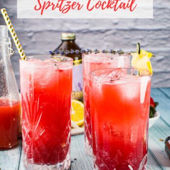 Three tall glasses of bright pink strawberry lavender spritzer cocktails with ice and garnishes sit on a light surface. A bottle, lemon slices, and sprigs of lavender are in the background. Text reads “Strawberry Lavender Spritzer Cocktail.”.