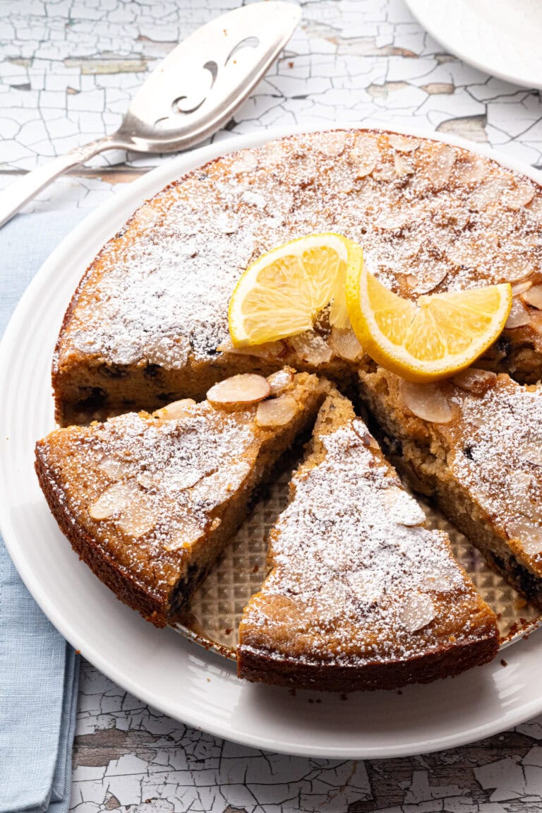 A gluten-free honey almond cake with wild blueberries, topped with powdered sugar and garnished with fresh lemon slices. One slice is cut and slightly pulled out, showing the moist interior with visible blueberries. The cake sits on a white plate over a woven placemat, with a cup of coffee, sliced lemons, and a jar of slivered almonds in the background. A mesh sifter with powdered sugar and a rustic wooden table add cozy, fall vibes.