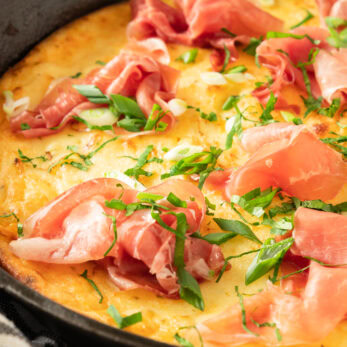 A close-up of a golden Gluten-Free Greek Yogurt Dutch Pancake , topped with thin slices of prosciutto, chopped green onions, and fresh herbs, with a striped towel nearby.