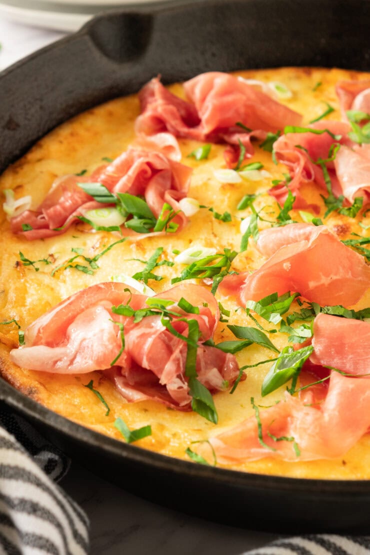 A close-up of a golden Gluten-Free Greek Yogurt Dutch Pancake , topped with thin slices of prosciutto, chopped green onions, and fresh herbs, with a striped towel nearby.