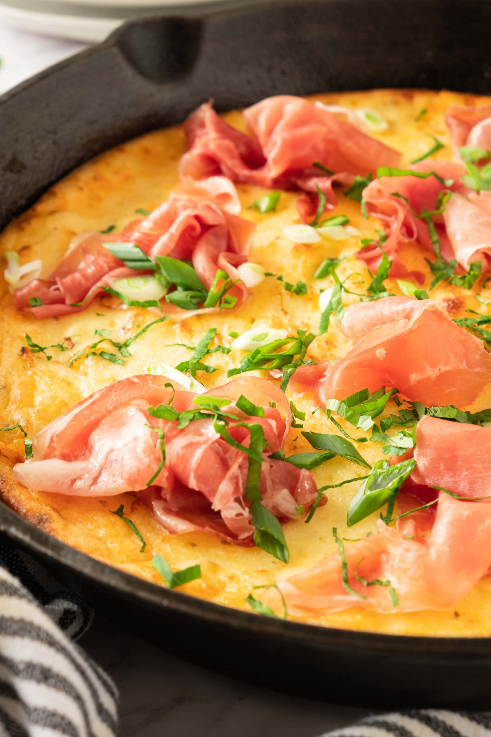 A close-up of a golden Gluten-Free Greek Yogurt Dutch Pancake , topped with thin slices of prosciutto, chopped green onions, and fresh herbs, with a striped towel nearby.