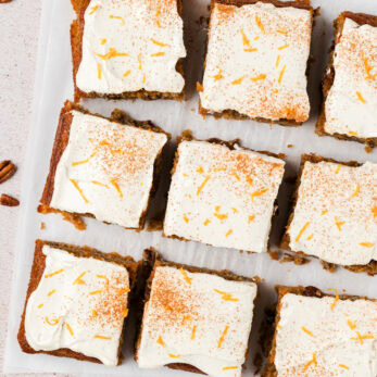 Nine square pieces of frosted carrot cake topped with cream cheese icing and orange zest. A cut orange, grated carrot, and pecans are visible at the edges of the image.