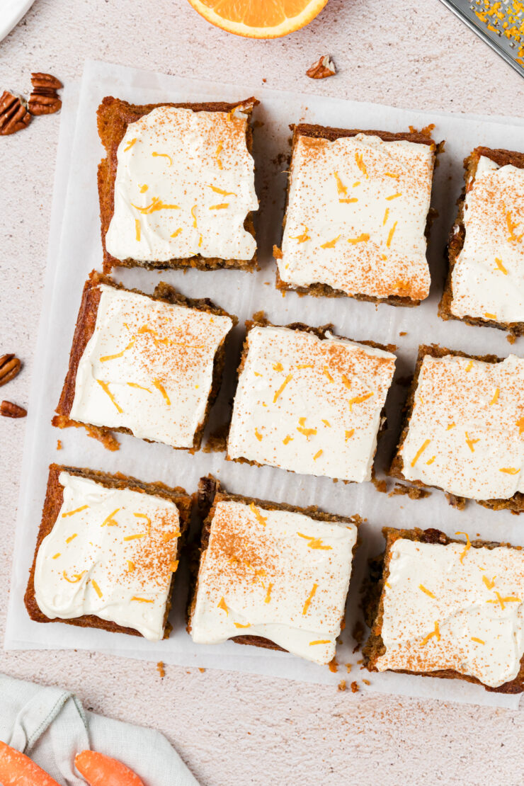 Nine square pieces of frosted carrot cake are arranged on parchment paper, topped with cream cheese icing and orange zest. A cut orange, grated carrot, and pecans are visible at the edges of the image.