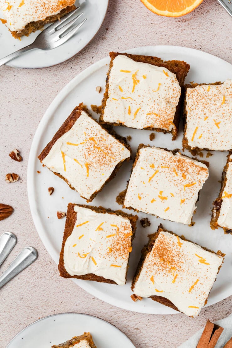 Top-down view of frosted cake squares on a white plate, garnished with orange zest and a sprinkle of cinnamon. Sliced oranges, pecans, and forks are visible around the plate on a light-colored surface.