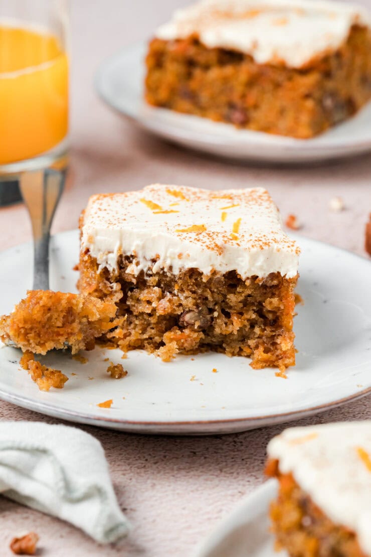 A square slice of carrot cake with white frosting sits on a plate, with a bite taken out. A fork rests nearby, and there is a glass of orange juice and more cake in the background.