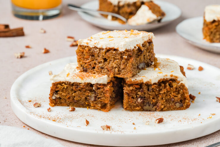 Three frosted carrot cake squares with chopped nuts are stacked on a white plate. More cake pieces are visible in the background, along with a glass of orange liquid and a fork on a plate.